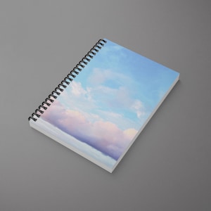 May include: A spiral bound notebook with a sky blue cover featuring a watercolour painting of clouds. The clouds are a light pink and white.