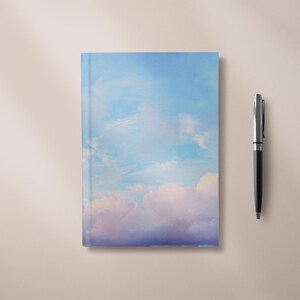 May include: A softcover notebook with a sky blue cover featuring a painting of fluffy white clouds against a light blue sky.