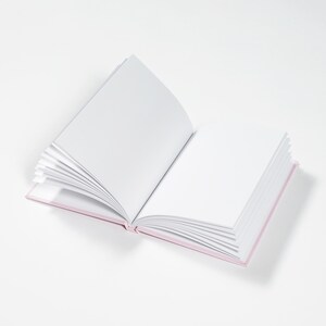May include: A blank, white book with a pink cover is open to a new page. The book is lying flat on a white surface.
