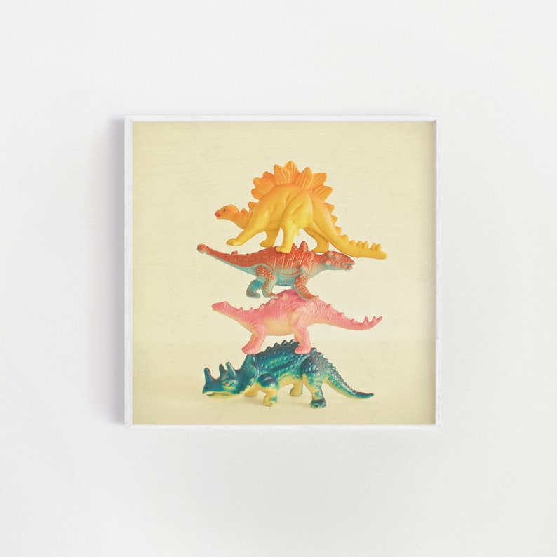 May include: A stack of four colorful plastic dinosaur toys. The dinosaurs are arranged from largest to smallest, with a yellow dinosaur on top, followed by an orange dinosaur, a pink dinosaur, and a blue dinosaur.