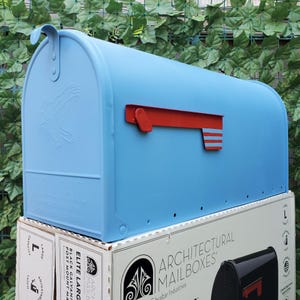 May include: A light blue, large capacity mailbox with a red flag and a decorative eagle emblem. The mailbox is sitting on top of a box labeled "Architectural Mailboxes".