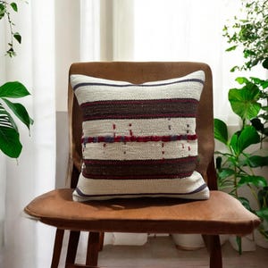 May include: A white, brown, and red striped throw pillow with a woven texture. The pillow is sitting on a brown leather chair.