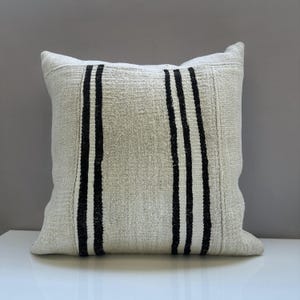 May include: Square decorative pillow with a textured, off-white fabric. The pillow features three vertical black stripes. The pillow is placed on a white surface.