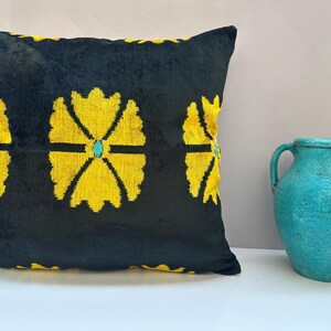 May include: A square black pillow with a repeating yellow floral pattern and turquoise accents. The pillow is next to a turquoise ceramic pitcher. The pillow is a decorative home accent.