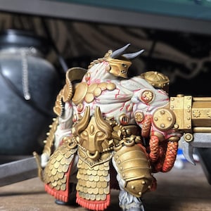 Op de afbeelding: Een wit en goud geverfd beeldje van een stierachtig wezen dat een harnas draagt en een geweer vasthoudt. Het wezen heeft rode accenten op zijn harnas en een gouden gezichtsmasker.