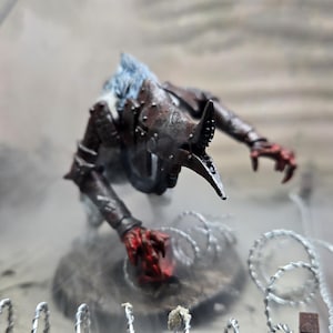 Puede incluir: Figura en miniatura detallada de una criatura con pico de ave y garras rojas, ambientada en un diorama de temática bélica con alambre de púas y terreno arenoso. La figura es de color marrón oscuro con pelo azul, la escena está envuelta en niebla.