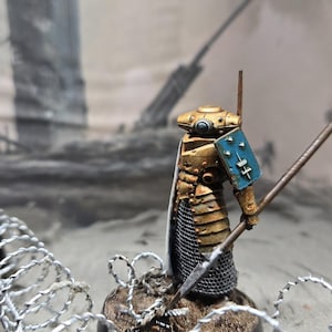 May include: A gold and silver painted miniature figure of a warrior holding a spear, standing on a round base with a brown and gray textured surface. The figure is wearing a helmet and armor with a blue and white shield. The figure is standing in front of a barbed wire fence.