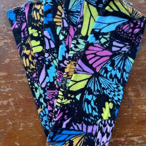 May include: A stack of colorful, patterned cloths featuring a butterfly design. The cloths have a black background with vibrant butterflies in shades of yellow, blue, pink, and purple. The edges are finished with a colorful stitch.