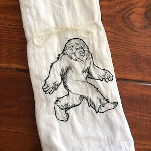 May include: White kitchen towel with a black embroidered image of Bigfoot walking. The towel is tied with a cream-colored string. The towel is made of a soft, textured fabric.