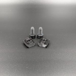 May include: Two black bolts and two black square nuts are arranged on a dark gray surface. The bolts have a threaded shaft and a circular head. The nuts have a square shape with a threaded hole in the center. The items appear to be hardware components.