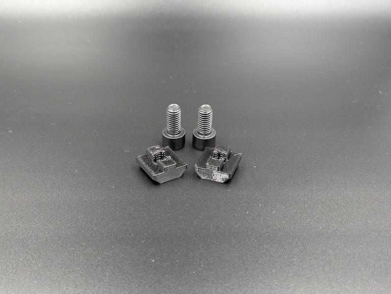 May include: Two black bolts and two black square nuts are arranged on a dark gray surface. The bolts have a threaded shaft and a circular head. The nuts have a square shape with a threaded hole in the center. The items appear to be hardware components.