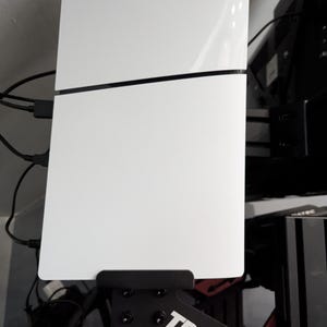 Può includere: Console di gioco bianca con una linea nera e il logo PlayStation. La console è montata su un supporto nero con la scritta "TRAK RAC" in bianco. Diversi cavi neri sono collegati alla console.