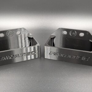 May include: Two black 3D-printed brackets with the word "FANATEC" embossed on the front. Each bracket has a rectangular base and a curved top with three holes. The brackets are designed to hold an item.