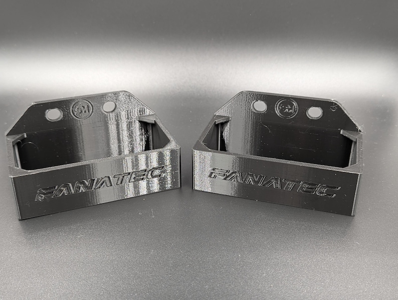 May include: Two black 3D-printed brackets with the word "FANATEC" embossed on the front. Each bracket has a rectangular base and a curved top with three holes. The brackets are designed to hold an item.