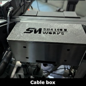May include: A black cable box with the text "SIM MOD WORKS" etched on the top. The box has several holes and a rectangular opening. This is a component of a racing simulator setup.