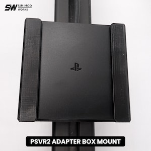Puede incluir: Un soporte de caja adaptadora PSVR2 negro que presenta una carcasa cuadrada central con el logotipo de PlayStation grabado en el centro. El soporte incluye dos rieles laterales verticales para una fijación segura, con el texto SIM MOD WORKS y PSVR2 ADAPTER BOX MOUNT visible.