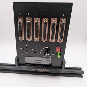 May include: A black vertical controller mount featuring the text SIM MOD WORKS at the top and THANOS at the base. The unit includes six DB25 ports numbered 1 through 6, a central rotary knob, directional arrow buttons, a reset button, and a small display.