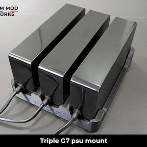 Op de afbeelding: Een zwarte Triple G7 psu-houder met drie rechthoekige voedingen. Elke eenheid heeft een glanzende afwerking en is aangesloten op een voedingskabel. De basis is donkergrijs. De tekst "Triple G7 psu mount" staat onderaan.