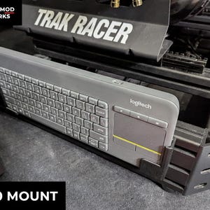 May include: A gray Logitech K400 keyboard with a built-in touchpad is mounted on a black metal bracket. The bracket is part of a larger black structure with the text "TRAK RACER" in white. The text "K400 MOUNT" is in a black box.