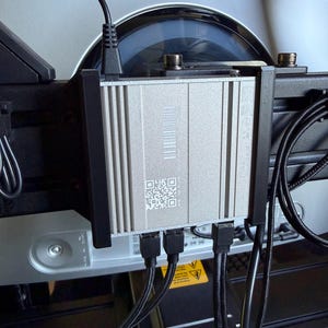 Peut inclure: Un appareil électronique argenté avec un code QR et un code-barres est monté sur un ordinateur. Des câbles noirs sont connectés à l'appareil. L'appareil est fixé à un écran d'ordinateur.