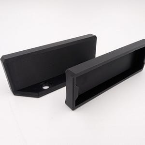 May include: Two black, geometric-shaped brackets. One bracket is rectangular with a recessed area, and the other is L-shaped with a hole. The brackets are made of a matte black material and are set against a white background.