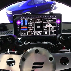 Puede incluir: Una configuración de simulador de carreras con un volante y una pantalla digital que muestra la velocidad, el tiempo por vuelta y otras métricas de rendimiento. El volante tiene levas de cambio y botones de control. Un coche azul es visible en el fondo.