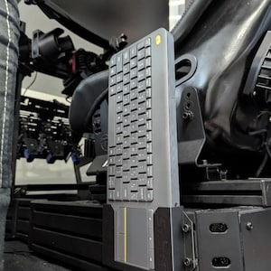 May include: A gray Logitech K400 Plus keyboard mounted on a black bracket. The keyboard has a built-in touchpad with a yellow accent. The text "LOGITECH K400 PLUS MOUNT" is displayed at the bottom of the image. The product is part of the Sim Mod Works product line.