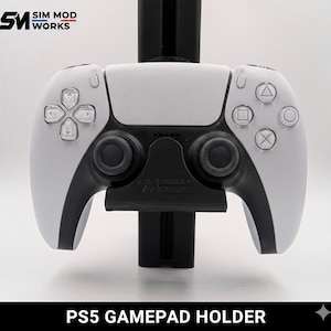 Puede incluir: Un mando de PS5 blanco y negro sostenido por un soporte negro. El mando tiene un diseño simétrico con botones y joysticks. La base del soporte tiene el texto "PS5 GAMEPAD HOLDER". El logotipo "SIM MOD WORKS" está en la esquina superior izquierda.