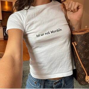 May include: White t-shirt with the text "lol ur not Wonbin" printed on the front. The shirt is being worn by a person with a brown leather handbag.