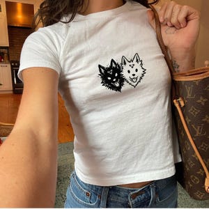 May include: A white short-sleeved t-shirt featuring a black graphic print of two stylized fox-like heads, one solid black and one white with black outlines, positioned centrally on the chest area of the garment, paired with blue denim jeans.