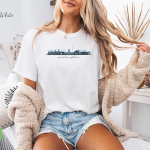 May include: White t-shirt featuring a skyline graphic of Washington, D.C. with the text "Washington" underneath. The model is wearing the shirt with denim shorts and a cream cardigan.
