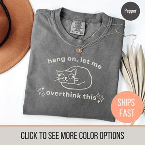May include: A grey Comfort Colors t-shirt with the text "hang on, let me overthink this" and a cat graphic. The shirt has a crew neck and short sleeves. A gold necklace is draped across the shirt.
