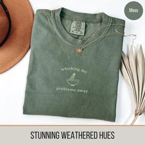 May include: A moss green t-shirt with the text "whisking my problems away" and a graphic of a mixing bowl with a whisk. The shirt is made of a soft, comfortable material and has a distressed look.