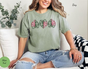 May include: A woman wearing a green t-shirt with a pink and green leaf design. The shirt has a distressed look and the leaves are in a row across the chest. The woman is sitting on a white couch with a black and white checkered throw blanket.