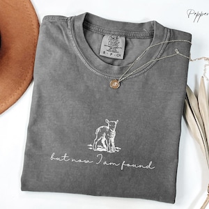 Puede incluir: Camiseta gris con un gráfico blanco de un cordero y el texto "but now I am found".
