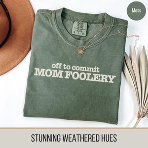 以下が含まれることがあります： 「off to commit MOM FOOLERY」の白い文字がプリントされたモスグリーンのTシャツ。金のネックレスとドライフラワーがシャツの横にあります。シャツには「Comfort Colors」とサイズS-P-CHのラベルが付いています。