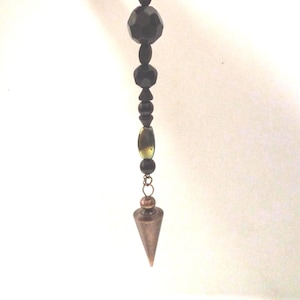 May include: A pendulum with a copper-colored cone-shaped weight. The pendulum chain features a series of black beads in various shapes and sizes, with a few olive-green beads near the top. The chain is suspended against a white background.