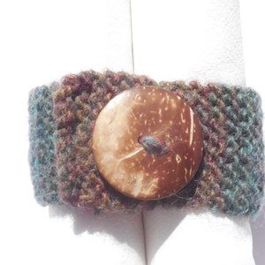 May include: A brown, green, and purple knitted wrist cuff with a large, round, natural coconut shell button.