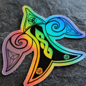 May include: A holographic sticker in a stylized leaf shape, featuring Celtic knot designs and symbols. The sticker has a rainbow color gradient, transitioning from purple to blue and yellow. Black outlines define the intricate patterns.