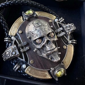 Puede incluir: Un conjunto de corbata bolo y pulsera de estilo steampunk. La corbata bolo presenta una calavera plateada sobre una base de madera, acentuada con piedras preciosas doradas y verdes. La pulsera tiene una letra "A" plateada con diamantes.