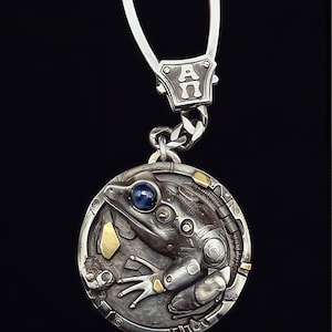 Puede incluir: Colgante de llavero plateado con un diseño detallado de rana, con un ojo de piedra preciosa azul. La rana está dentro de un marco circular con detalles mecánicos y detalles dorados. El llavero tiene una cadena plateada y un cierre con las letras "A" y "N".
