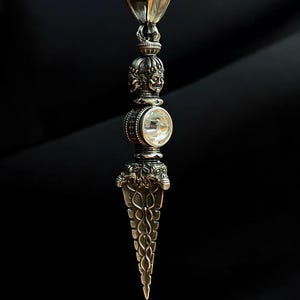 May include: A silver pendant with an intricate design. It features a clear, round stone and a decorative, pointed base. The top has a heart-shaped bail and a skull-like figure. The pendant hangs from a dark brown leather cord. The text "BUDDHIST collection" is in the lower left corner.