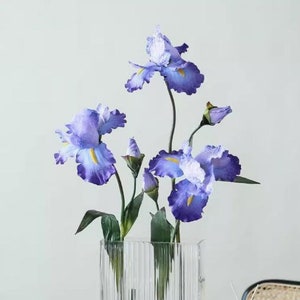 May include: A bouquet of purple iris flowers in a clear glass vase with vertical lines. The vase is on a table with a stack of books that say "Arts the Crafts" on the cover.