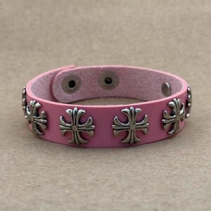 May include: A pink leather bracelet featuring a series of evenly spaced silver-toned metal cross emblems. The bracelet includes two circular metal snap fasteners on the interior side for closure, providing a secure fit around the wrist.