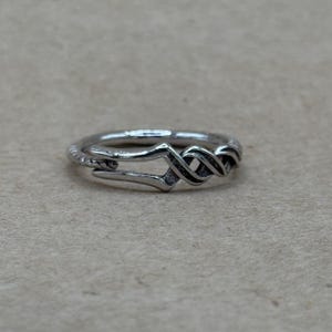 May include: A silver-toned ring featuring a polished band that transitions into an intricate, twisted knot design at the front. The metal has a slightly oxidized finish in the recessed areas, highlighting the detailed, interwoven metalwork.