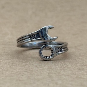 May include: A silver-toned metal ring shaped like an open-ended wrench that wraps around the finger. The band features a textured, industrial design with a wrench head on one end and a box-end wrench head on the other, creating a continuous loop.