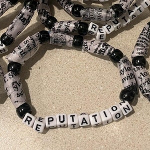 K&ouml;nnte beinhalten: Nahaufnahme von mehreren schwarzen und wei&szlig;en Perlenarmb&auml;ndern, auf denen jeweils das Wort "REPUTATION" steht.