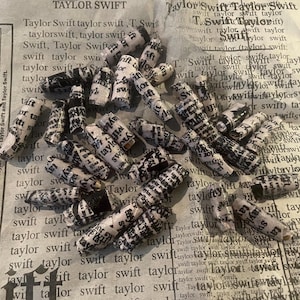 K&ouml;nnte beinhalten: Eine Sammlung kleiner, wei&szlig;er Papierrollen mit schwarzem Text darauf. Die Rollen sind aufgerollt und haben eine leicht strukturierte Oberfl&auml;che. Der Text auf den Rollen ist haupts&auml;chlich der Name "Taylor Swift", der viele Male wiederholt wird.
