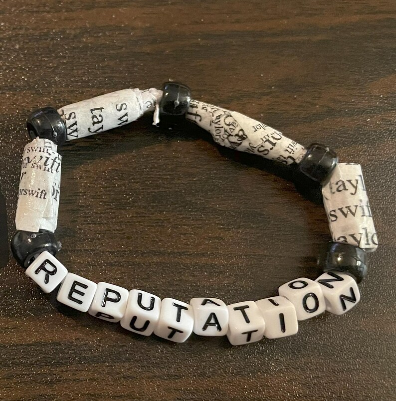 K&ouml;nnte beinhalten: Ein wei&szlig;es Perlenarmband mit schwarzen Perlen und dem Wort "REPUTATION" auf den wei&szlig;en Perlen.