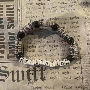 K&ouml;nnte beinhalten: Ein schwarz-wei&szlig;es Perlenarmband mit dem Wort "REPUTATION" in wei&szlig;en Perlen. Die Perlen bestehen aus Zeitungsartikeln mit dem Wort "Taylor Swift" darauf.